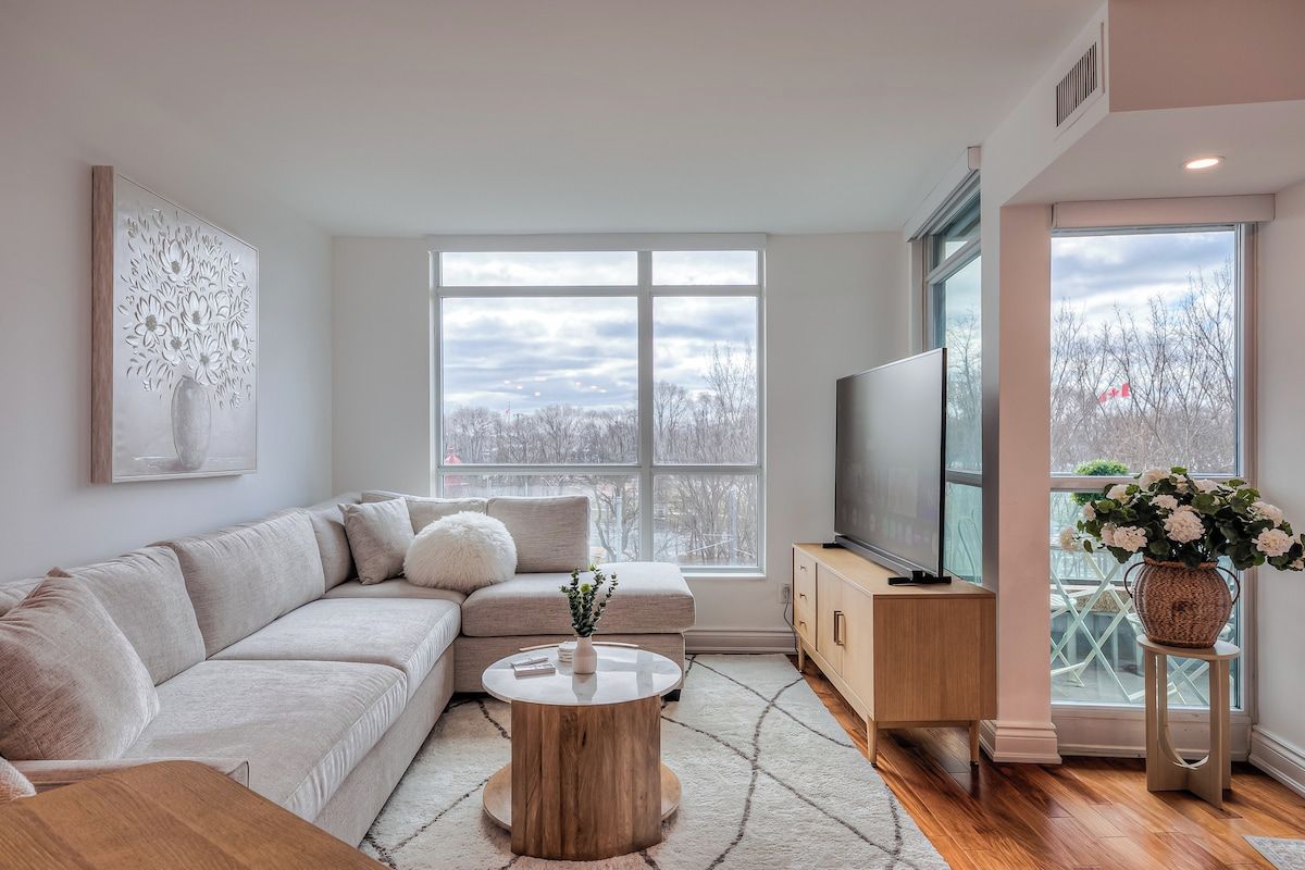 A modern living room with a sectional sofa, a round coffee table, a flat-screen TV, and a large window offering a view of trees.