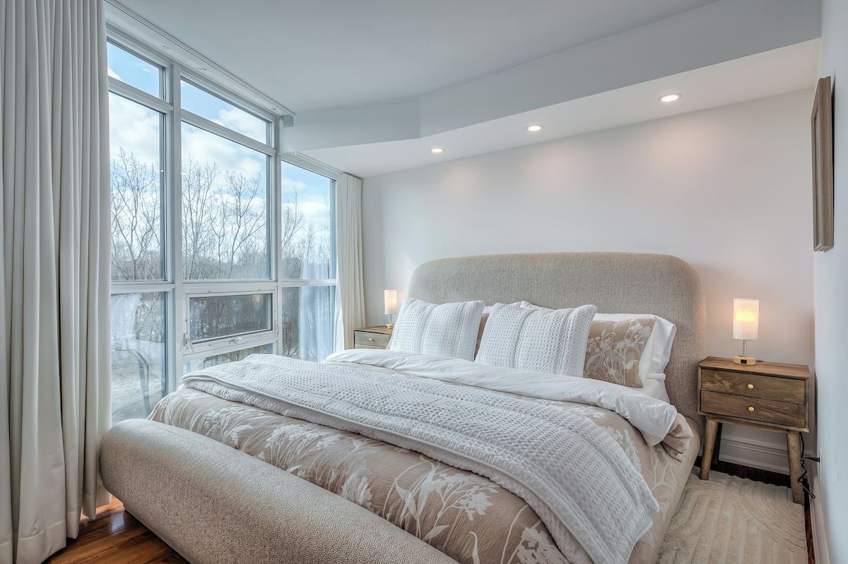 Un dormitorio con un ventanal grande, una cama con ropa de cama blanca, una mesita de noche con una lámpara y suelo de madera.