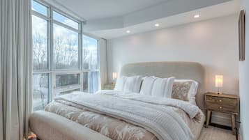 Un dormitorio con un ventanal grande, una cama con ropa de cama blanca, una mesita de noche con una lámpara y suelo de madera.