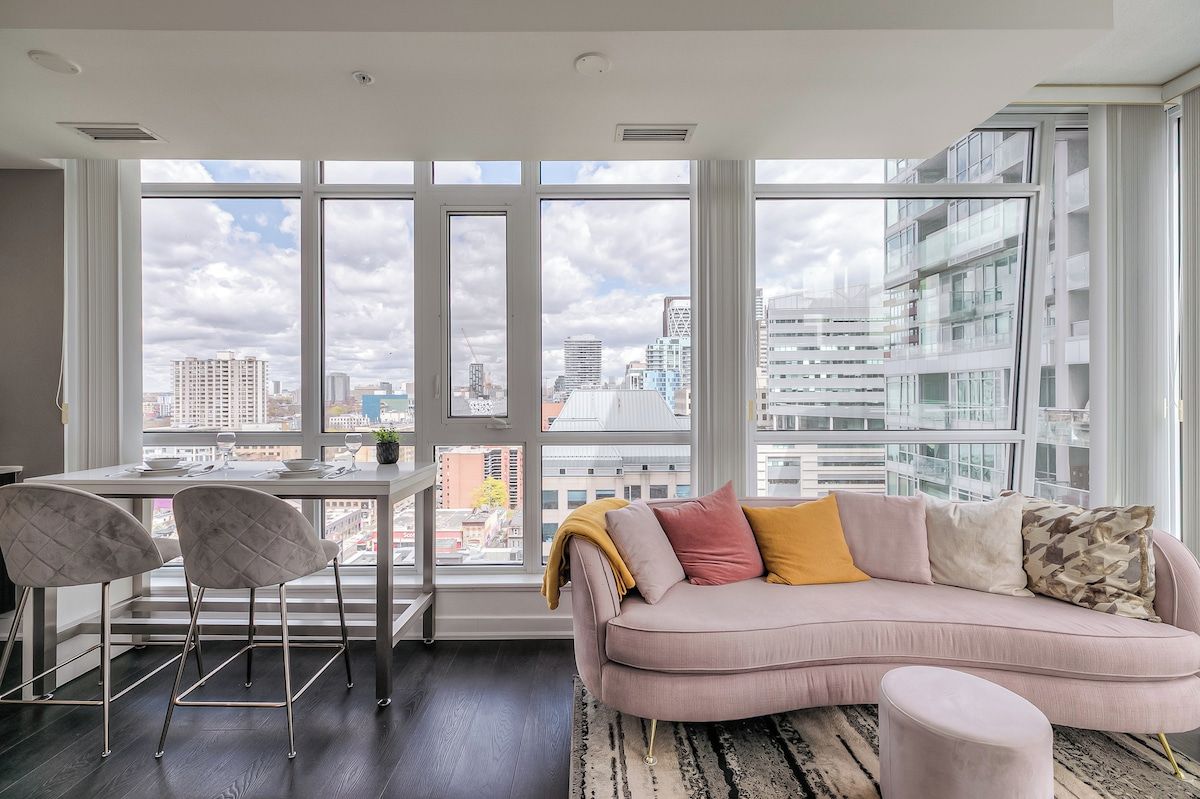 Uma sala de estar moderna com uma janela grande, um sofá, uma mesa de jantar com cadeiras e vista para a paisagem urbana.