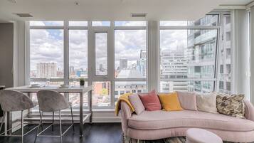 Uma sala de estar moderna com uma janela grande, um sofá, uma mesa de jantar com cadeiras e vista para a paisagem urbana.