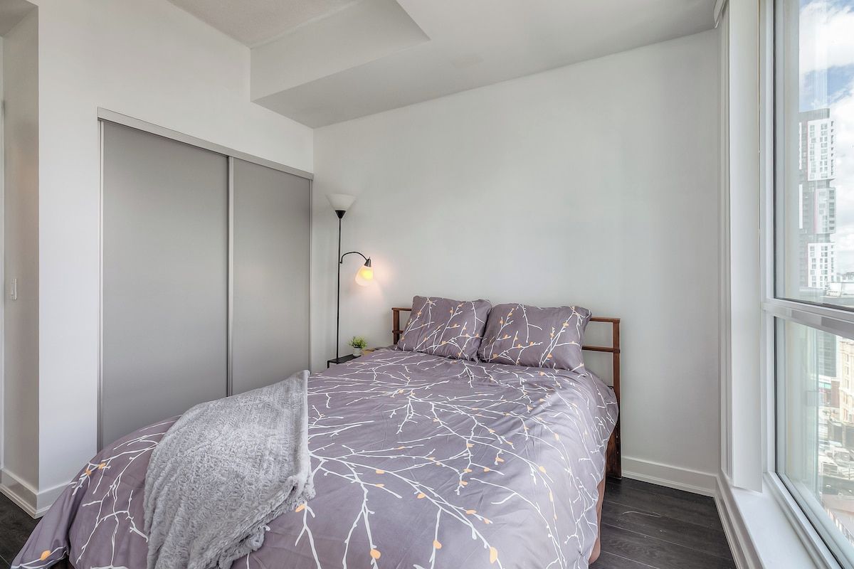 Une chambre à coucher avec un lit, une table de chevet, une lampe et une fenêtre donnant sur des immeubles.