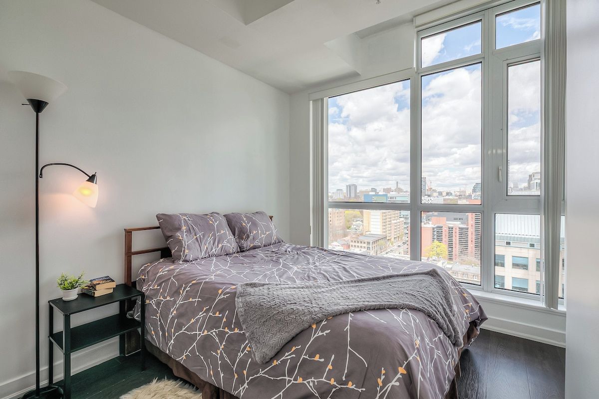 A bedroom with a large window, a bed with a patterned bedspread, a bedside table with a lamp, and a view of the cityscape.