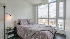 Une chambre avec une grande fenêtre, un lit recouvert d’une couvre-lit à motifs, une table de chevet avec une lampe, et une vue sur le paysage urbain.