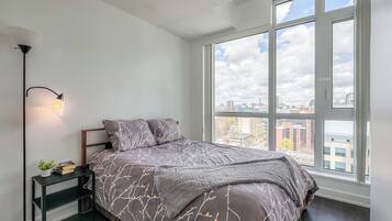 A bedroom with a large window, a bed with a patterned bedspread, a bedside table with a lamp, and a view of the cityscape.