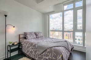 A bedroom with a large window, a bed with a patterned bedspread, a bedside table with a lamp, and a view of the cityscape. - CN Tower Views - 2BR Condo - Balcony - Fast Wi-Fi (Toronto)