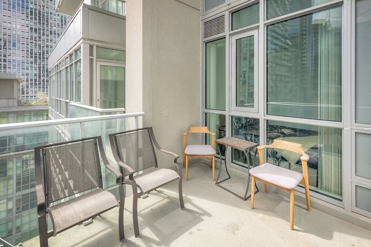 Varanda com duas cadeiras de metal e uma de madeira, uma mesa pequena e vista para a cidade através de grandes janelas.