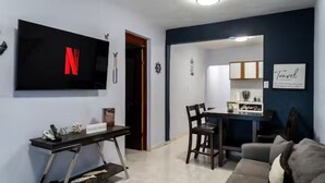 Ein Wohnzimmer mit Fernseher, einem Essbereich mit Stühlen, einer Couch und einer Küchenzeile mit Schränken.