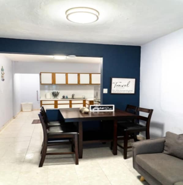 A dining area with a dark wooden table and chairs, a kitchen in the background, and a wall with a framed sign. - Charming 2BR in Aguada, PR, with AC Relax & Unwind (Aguada)