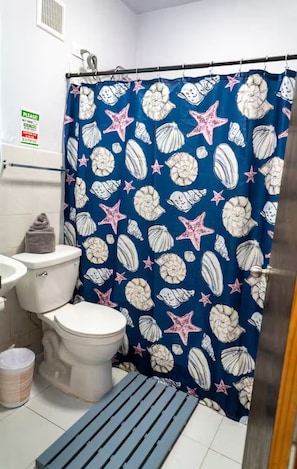 A bathroom with a blue shower curtain featuring a seashell and starfish pattern, a white toilet, and a small sink. - Charming 2BR in Aguada, PR, with AC Relax & Unwind (Aguada)