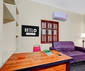 A hotel room with a wooden table, a purple sofa, a sign indicating it's serviced by OYO, and a window with a metal grille. - Classic With Balcony Rama Home Stay (Tirupati)