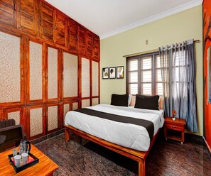A hotel room with a large bed, wooden wardrobe, a small bedside table, a window with curtains, and a tiled floor.
