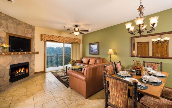 A living room with a fireplace, a television, a sofa, a dining table set for four, and a balcony with a view.