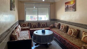 A room with a sofa set, a round table, and a window with blinds. - Cozy Urban Tanger 03 (Tanger)