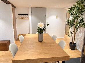 Une salle de conférence avec une longue table en bois, des chaises blanches, un vase avec des fleurs, un porte-manteau et une plante en pot.