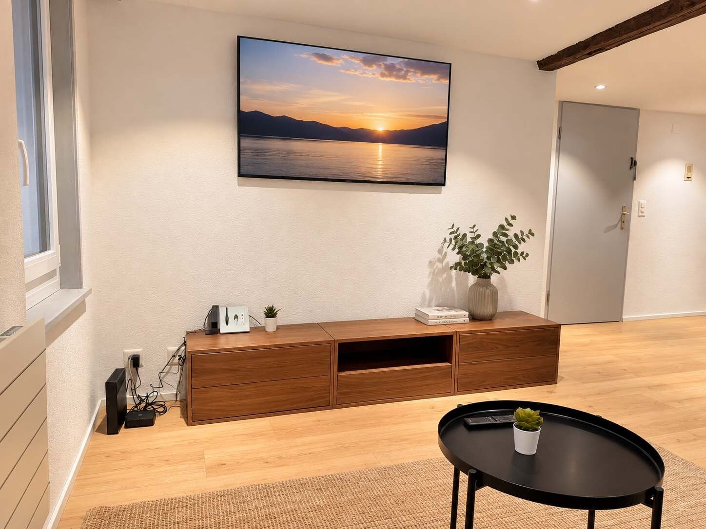 A room with a wooden entertainment unit, a black table with a plant, and a wall-mounted TV displaying a sunset over water.