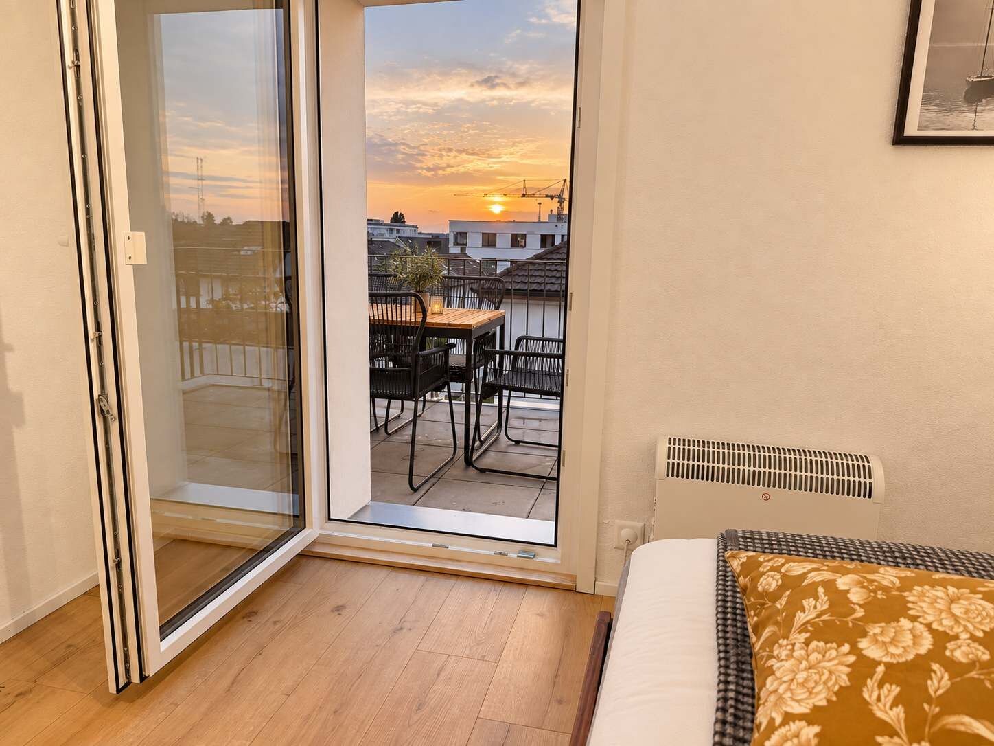 Une chambre avec une porte-fenêtre coulissante donnant sur un balcon équipé d’une table et de chaises, un lit agrémenté d’un coussin à motifs et une grille d’aération au mur.