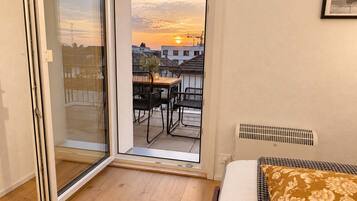 Une chambre avec une porte-fenêtre coulissante donnant sur un balcon équipé d’une table et de chaises, un lit agrémenté d’un coussin à motifs et une grille d’aération au mur.