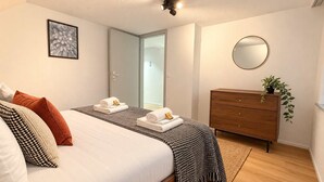 A modern bedroom with a bed, bedside tables, a dresser, a mirror, and a door leading to a bathroom. - book2stay: Apartment with 2 bedrooms, balcony near harbor & train station (Romanshorn)