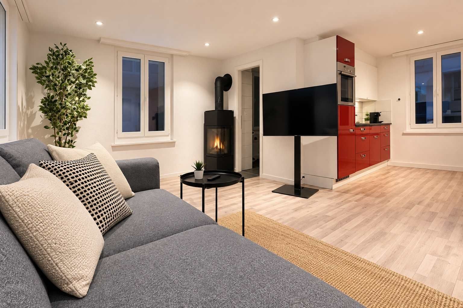 Un soggiorno moderno con un divano grigio, una TV a schermo piatto, un camino e un angolo cucina rosso.