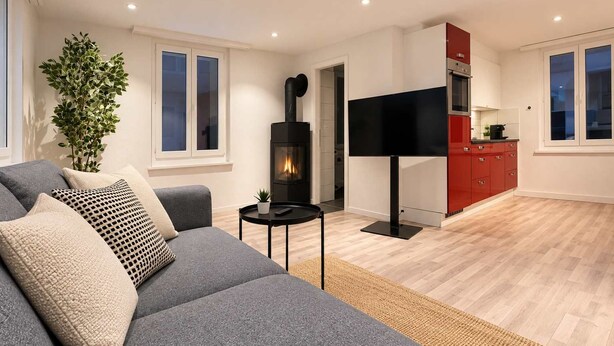 A modern living room with a grey sofa, a flat-screen TV, a fireplace, and a red kitchen area. - book2stay: Apartment with Balcony near harbor & train station (Romanshorn)