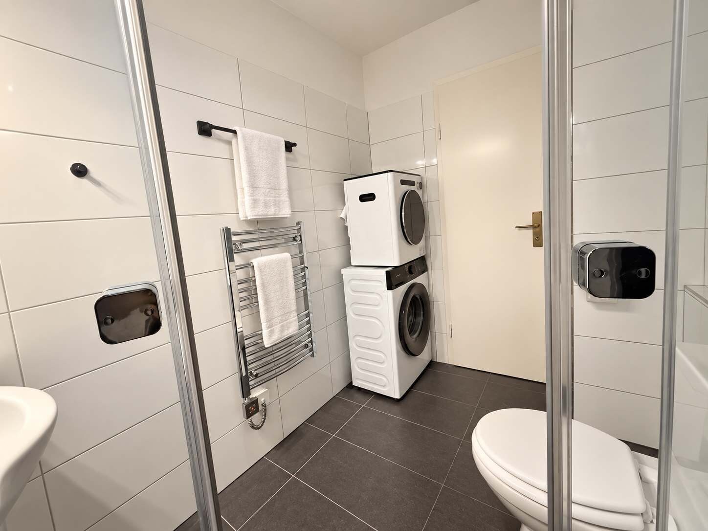 Une salle de bain moderne avec une douche à parois de verre, un lave-linge et un sèche-linge superposés, et des toilettes.