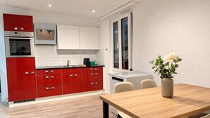 A modern kitchen with red cabinetry, a built-in oven, and a dining area with a wooden table and chairs. - book2stay: Apartment with Balcony near harbor & train station (Romanshorn)
