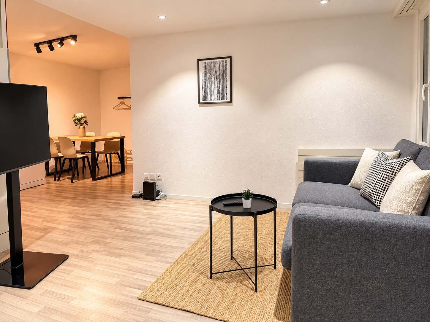 A modern living room with a flat-screen TV, a grey sofa, a small round table, and a dining area in the background.