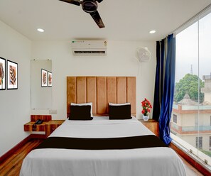 A hotel room with a large bed, a ceiling fan, an air conditioning unit, a bedside table with a phone, a mirror, and a window with a view of buildings and trees. - Deluxe Govindpuri Station Formerly Montto Plaza (Kanpur)