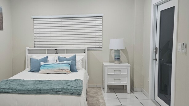 Une chambre à coucher avec un lit, une table de chevet avec une lampe, une fenêtre avec des stores et une porte.