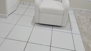 Une pièce avec un sol carrelé de carreaux blancs, un fauteuil blanc, une armoire blanche à portes à persiennes et un mur blanc.