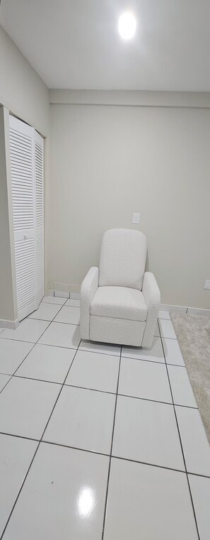 Um quarto com piso de azulejos brancos, uma poltrona branca, um guarda-roupa branco com portas venezianas e uma parede branca.