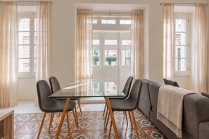 Espaço de refeições com mesa de vidro e cadeiras, sofá cinza e ladrilhos com padrão.