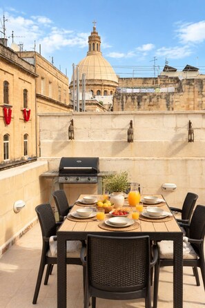 An outdoor dining area with a grill, table set with plates, glasses, and a pitcher of orange juice. - Historic Valletta House, 3BR & Terrace (Valletta)