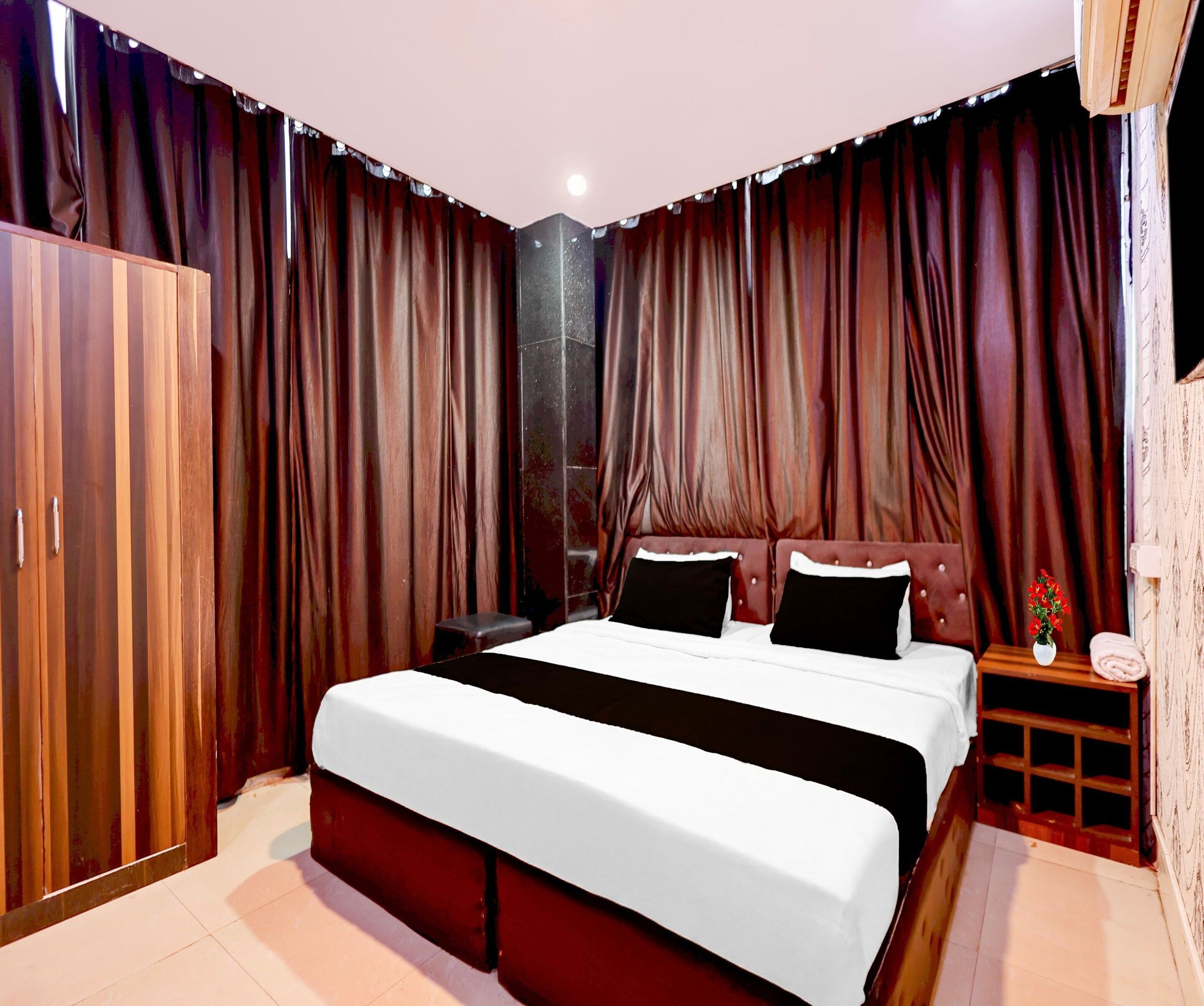 A bedroom with a large bed, a wardrobe, a nightstand with a lamp, and a window with curtains.