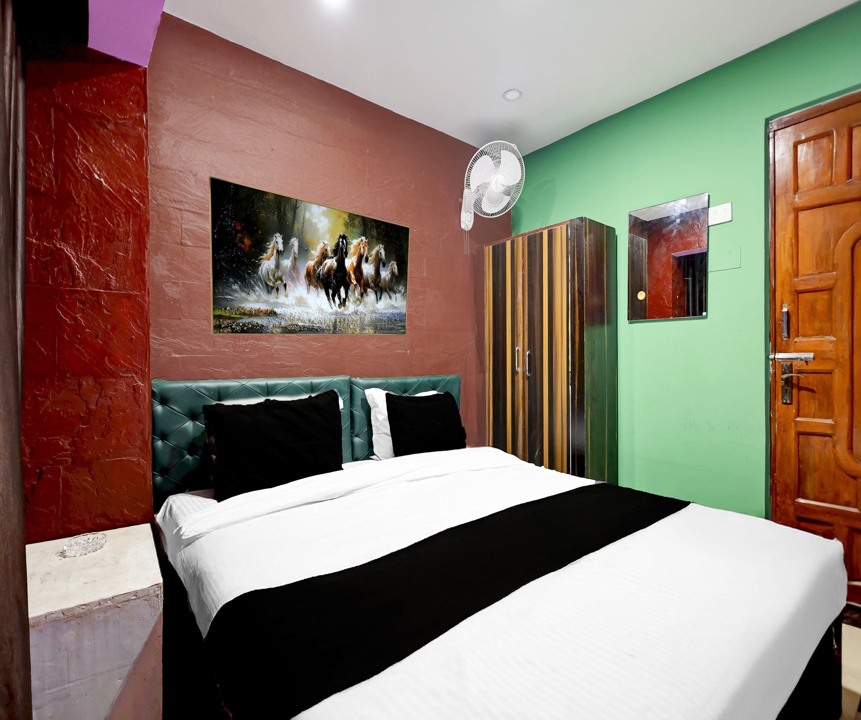 Una camera d'albergo con un letto, un quadro raffigurante cavalli, una ventola e un armadio.
