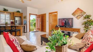A modern living room with a sofa, coffee table, and a television. There is a kitchen area with a refrigerator and a water dispenser. The room has a ceiling fan and a door leading outside.