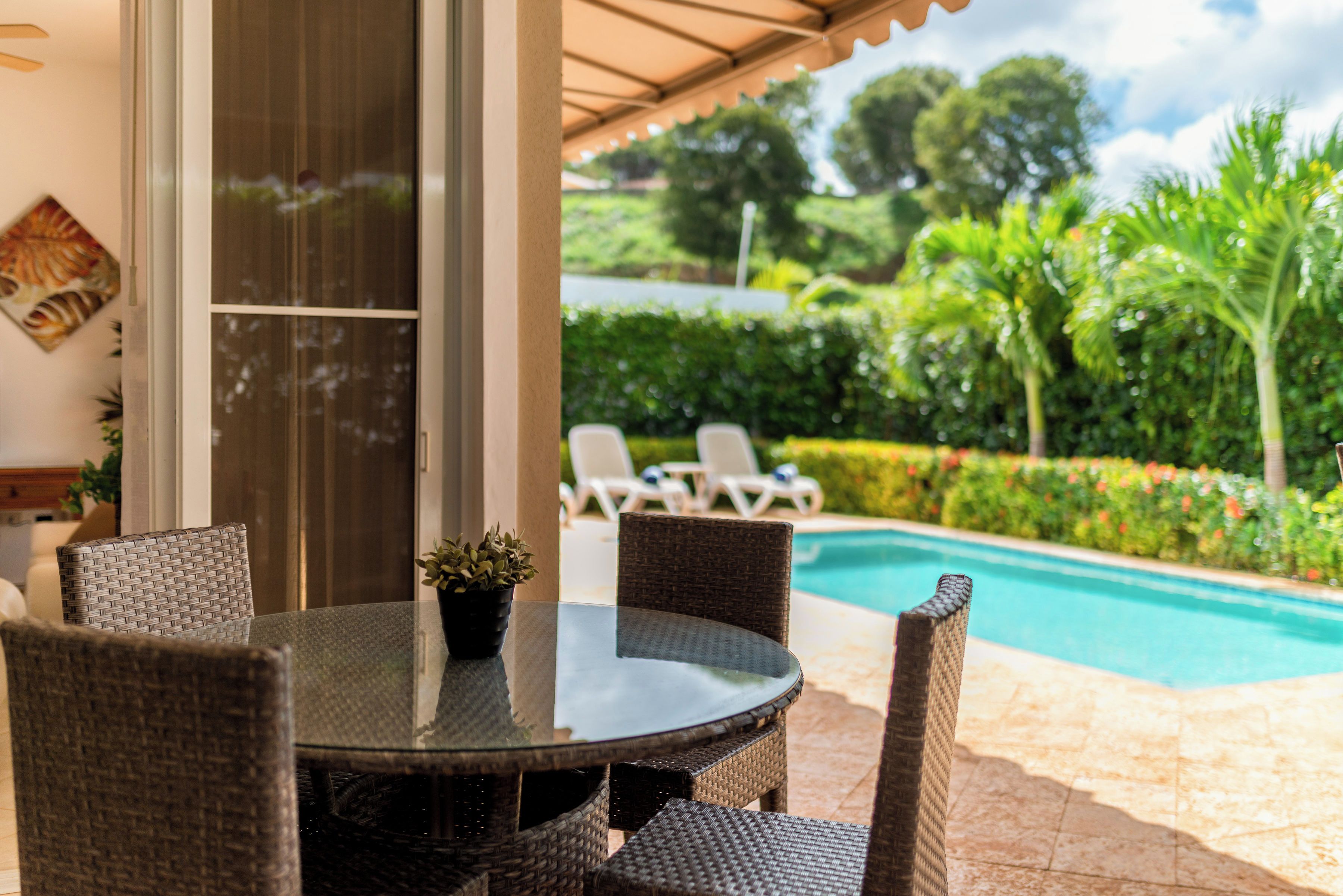 Un patio con muebles de mimbre, una mesa de vidrio y vistas a una piscina y follaje.