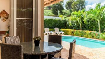Un patio con muebles de mimbre, una mesa de vidrio y vistas a una piscina y follaje.