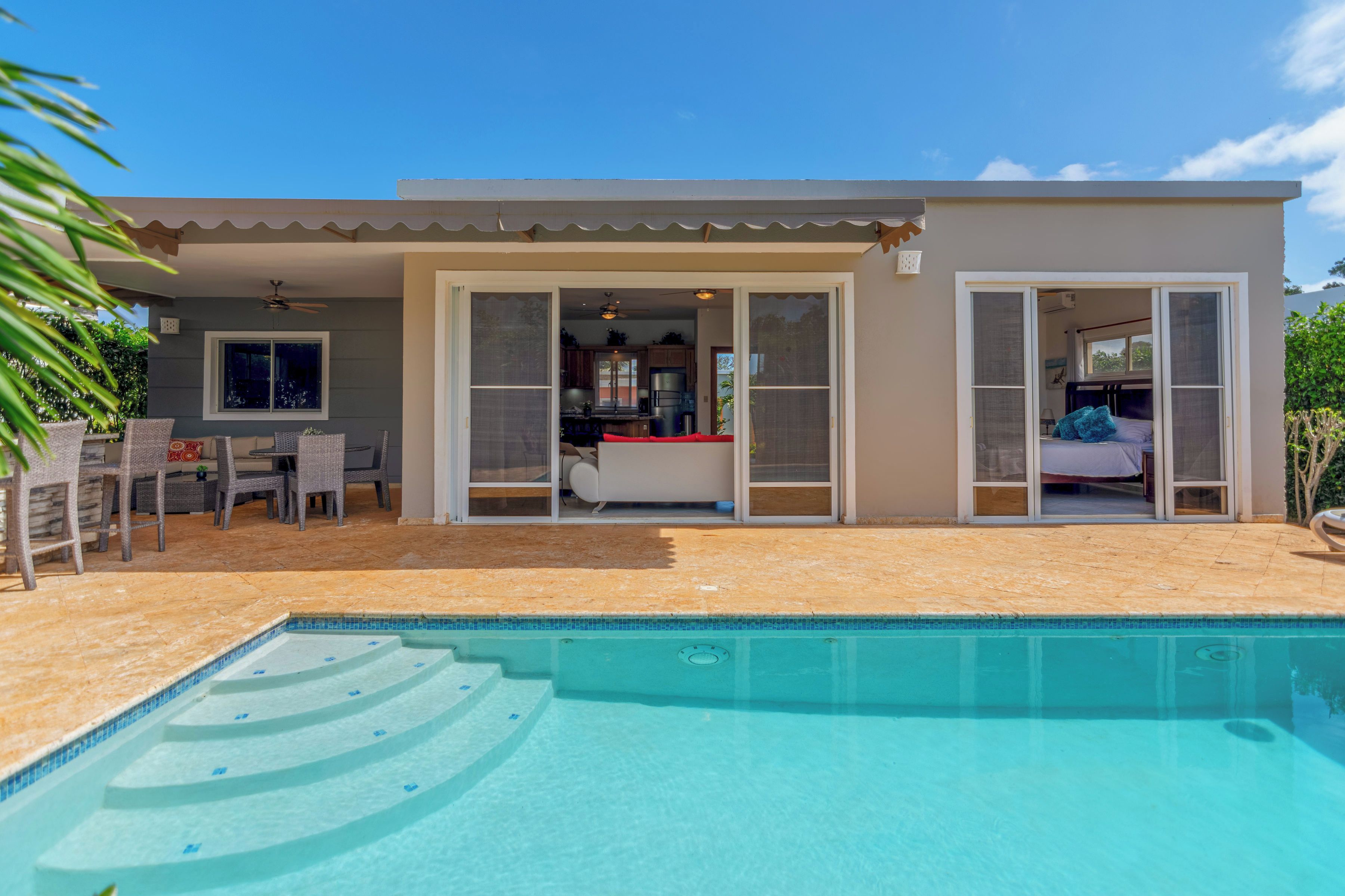 A modern house with a swimming pool, a patio with outdoor furniture, and a large sliding glass door leading to a kitchen.