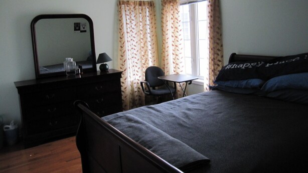 A bedroom with a bed, a chair, a small table, a dresser, and a window with curtains. - Cosy Room with exclusive bathroom (Mississauga)