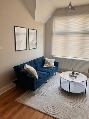 A modern living room with a blue sofa, two framed artworks, a round coffee table, and a large window with blinds. - Modern Apartment | 5 min Walk to Watford Junction (Watford)
