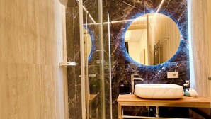 Ein modernes Badezimmer mit einer gläsernen Duschkabine, einer Wand mit Marmorplatten, einem runden Spiegel und einem Holz-Waschtisch.