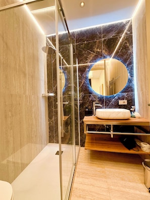 Un baño moderno con ducha envuelta en cristal, pared de azulejos de mármol, espejo redondo y tocador de madera.