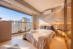 A modern bedroom with a large window, a bed, a bedside table, and a wall-mounted air conditioning unit. - Yuny - Zip  Morumbi (São Paulo)