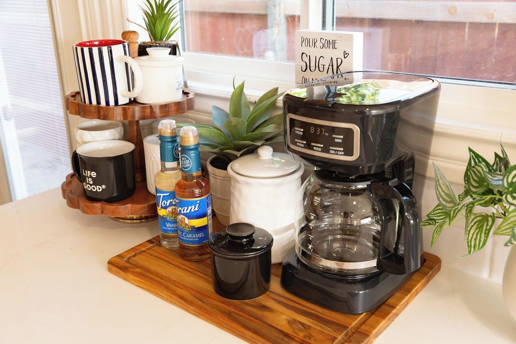 A coffee maker on a wooden tray with various mugs, a sign, and condiments.