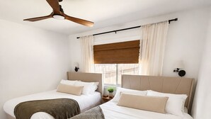 Un dormitorio con dos camas, un ventilador de techo, una ventana con cortinas y suelo de madera.