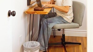 Uma pessoa sentada em uma escrivaninha de madeira, com um laptop, em um quarto com uma porta branca, uma lixeira e um objeto decorativo fixado na parede.