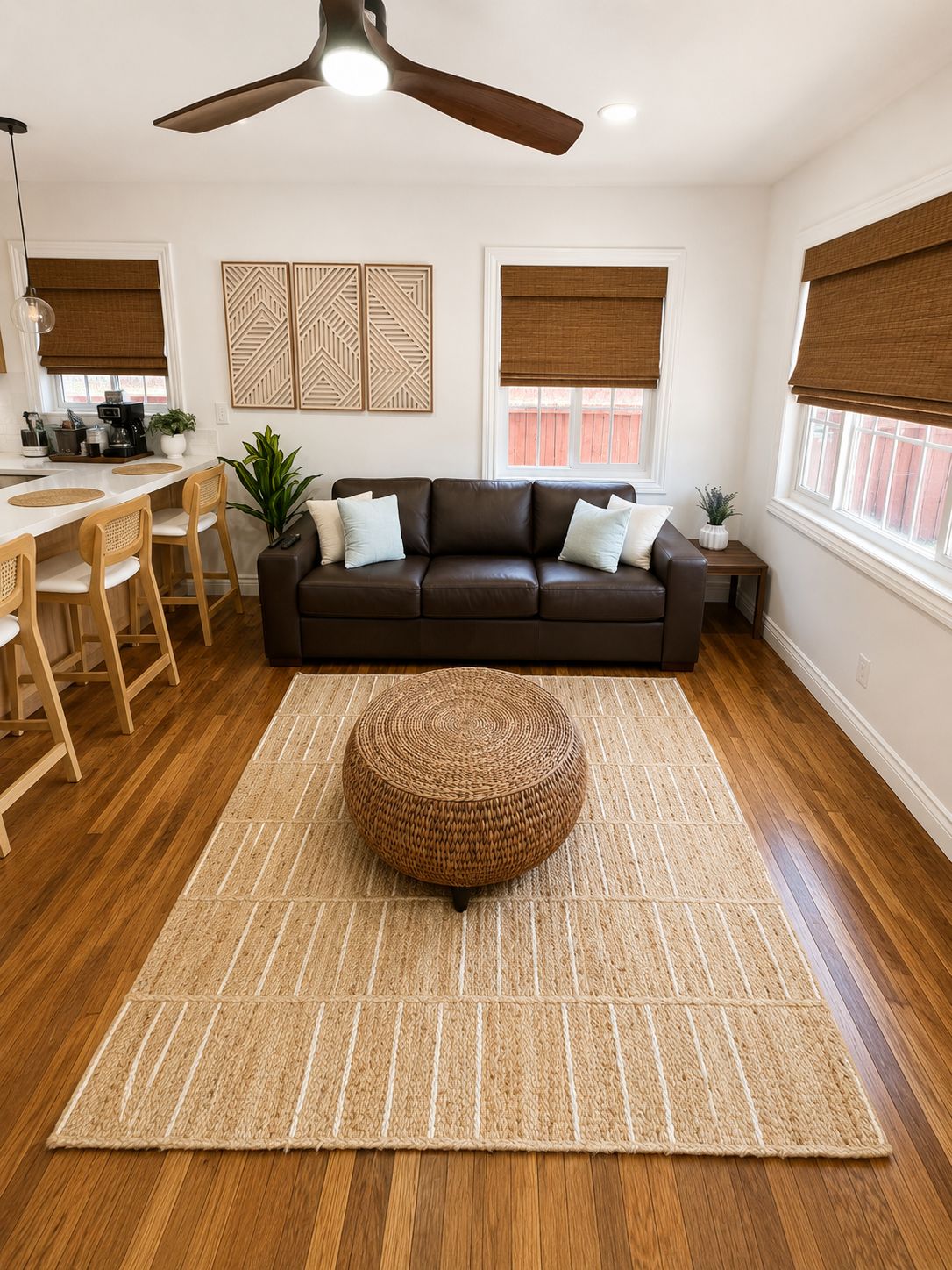 A modern living room with a brown leather sofa, a round woven ottoman, a dining area with wooden chairs, a kitchen with a white countertop, and a ceiling fan.
