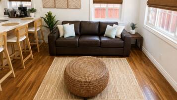 A modern living room with a brown leather sofa, a round woven ottoman, a dining area with wooden chairs, a kitchen with a white countertop, and a ceiling fan.